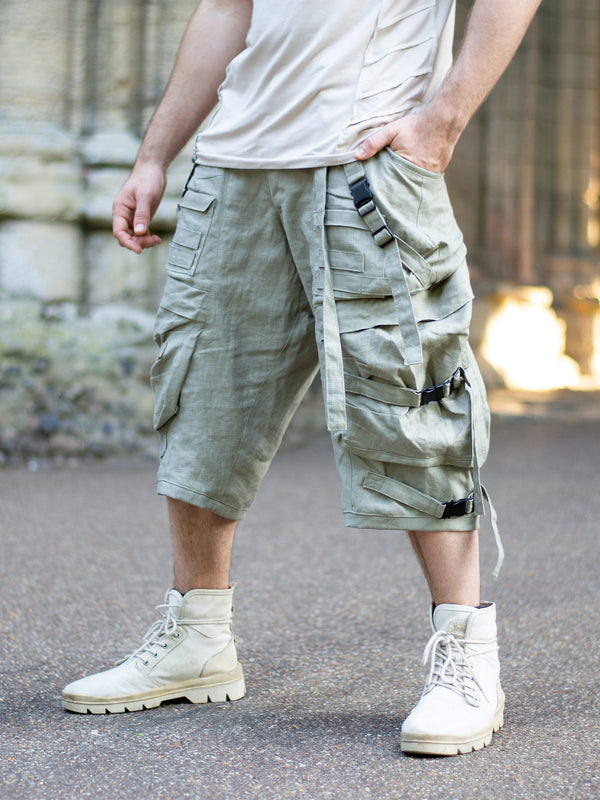 Men’s sage green avant-garde linen cargo shorts with drop crotch silhouette, 10 functional pockets, metal buckles, tactical straps, and below-knee cut. Techwear-style festival shorts designed for breathability and movement.