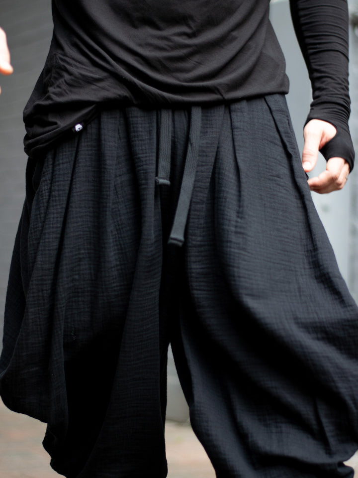 Model wearing black drop-crotch ninja pants inspired by traditional shinobi attire — Hattori harem pants in breathable muslin with pleated volume, calf supports, and a flowy, avant-garde silhouette.