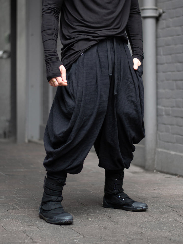Model wearing black drop-crotch ninja pants inspired by traditional shinobi attire — Hattori harem pants in breathable muslin with pleated volume, calf supports, and a flowy, avant-garde silhouette.