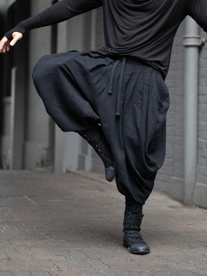 Model wearing black drop-crotch ninja pants inspired by traditional shinobi attire — Hattori harem pants in breathable muslin with pleated volume, calf supports, and a flowy, avant-garde silhouette.