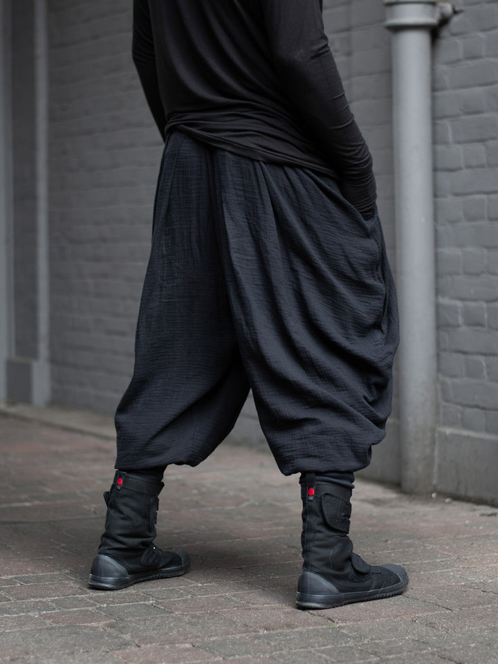 Model wearing black drop-crotch ninja pants inspired by traditional shinobi attire — Hattori harem pants in breathable muslin with pleated volume, calf supports, and a flowy, avant-garde silhouette.