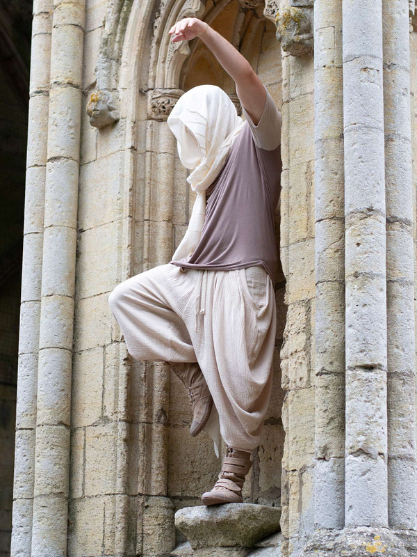 Sand drop-crotch ninja style pants made from lightweight double gauze muslin cotton, featuring wide pleated legs, ribbed calf supports, and a flowy silhouette. Inspired by traditional Japanese attire with an avant-garde, streetwear twist — ideal for summer layering, movement, and alternative festival fits.