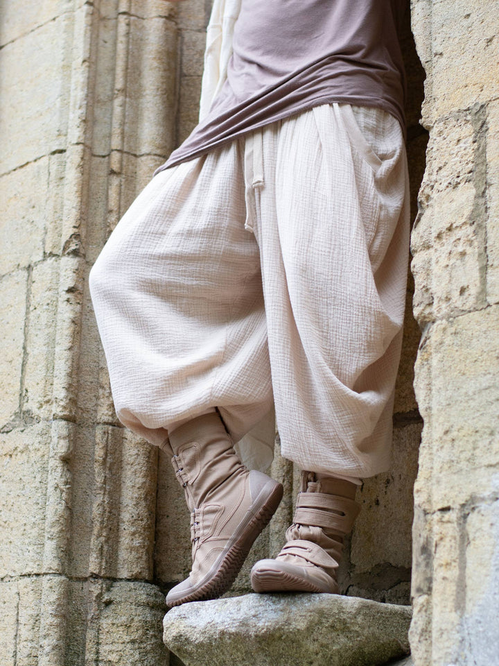 Sand drop-crotch ninja style pants made from lightweight double gauze muslin cotton, featuring wide pleated legs, ribbed calf supports, and a flowy silhouette. Inspired by traditional Japanese attire with an avant-garde, streetwear twist — ideal for summer layering, movement, and alternative festival fits.