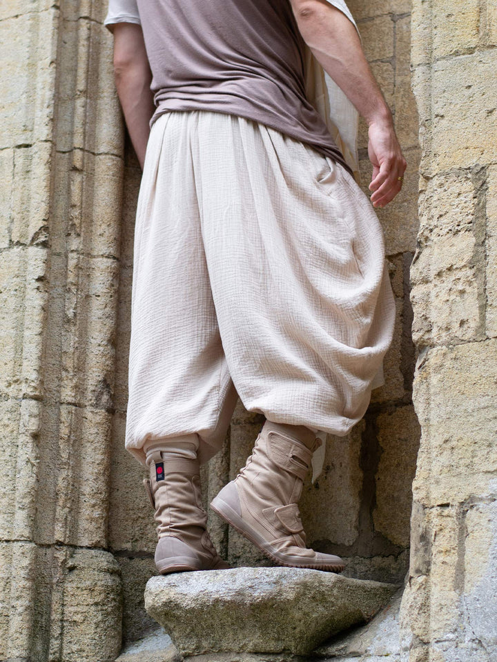 Sand drop-crotch ninja style pants made from lightweight double gauze muslin cotton, featuring wide pleated legs, ribbed calf supports, and a flowy silhouette. Inspired by traditional Japanese attire with an avant-garde, streetwear twist — ideal for summer layering, movement, and alternative festival fits.