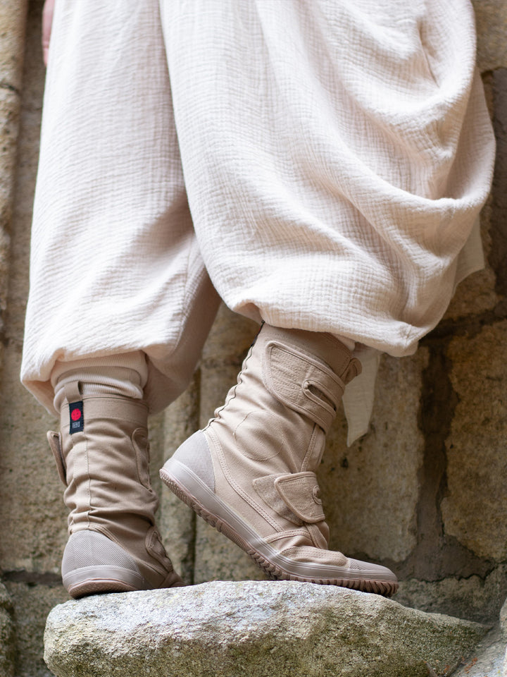 Sand drop-crotch ninja style pants made from lightweight double gauze muslin cotton, featuring wide pleated legs, ribbed calf supports, and a flowy silhouette. Inspired by traditional Japanese attire with an avant-garde, streetwear twist — ideal for summer layering, movement, and alternative festival fits.