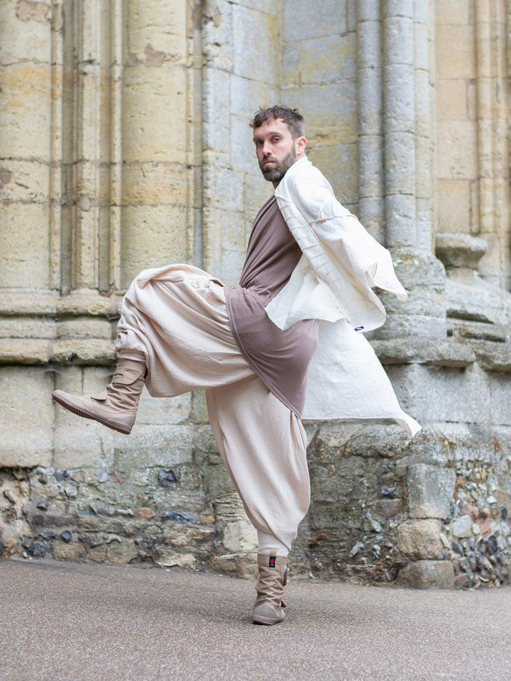 Sand drop-crotch ninja style pants made from lightweight double gauze muslin cotton, featuring wide pleated legs, ribbed calf supports, and a flowy silhouette. Inspired by traditional Japanese attire with an avant-garde, streetwear twist — ideal for summer layering, movement, and alternative festival fits.
