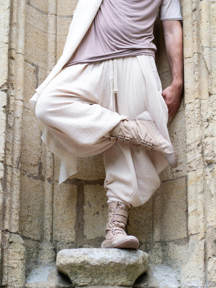 Sand drop-crotch ninja style pants made from lightweight double gauze muslin cotton, featuring wide pleated legs, ribbed calf supports, and a flowy silhouette. Inspired by traditional Japanese attire with an avant-garde, streetwear twist — ideal for summer layering, movement, and alternative festival fits.