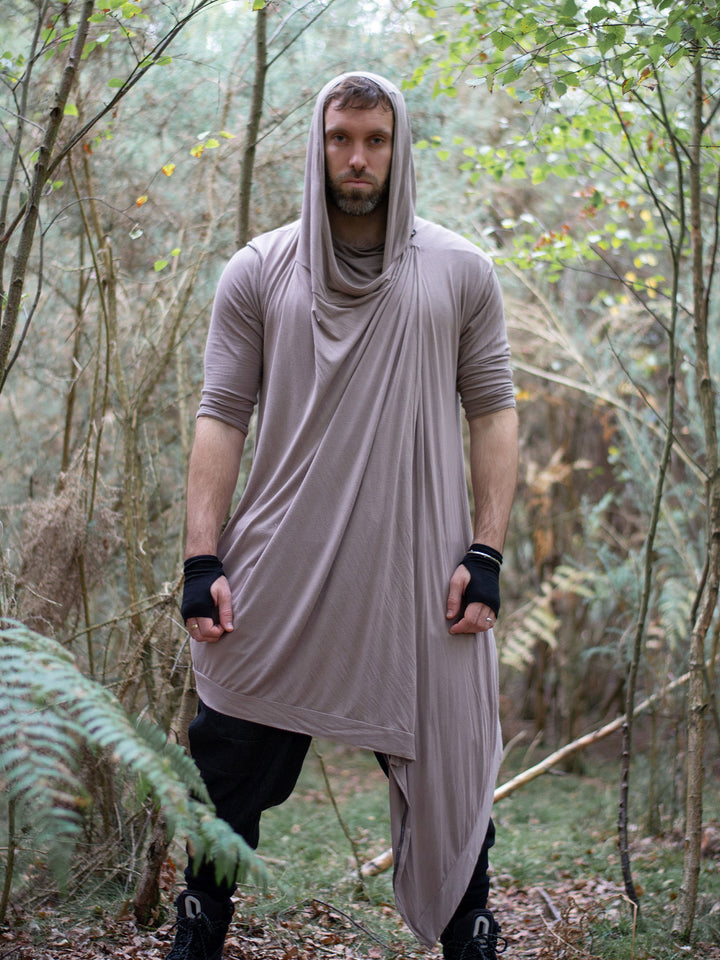 Men’s avant garde hooded cloak in sand with asymmetric wrap-over cascade design. Features bamboo jersey fabric, oversized hood, and wooden shoulder button for layered post-apocalyptic styling.