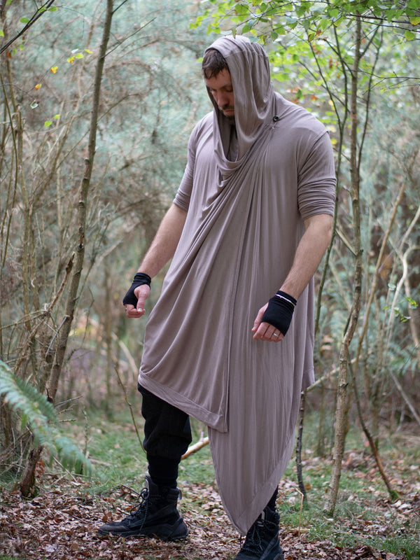 Men’s avant garde hooded cloak in sand with asymmetric wrap-over cascade design. Features bamboo jersey fabric, oversized hood, and wooden shoulder button for layered post-apocalyptic styling.