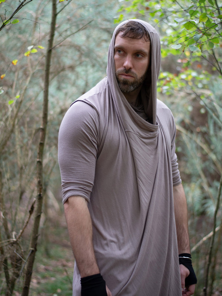 Men’s avant garde hooded cloak in sand with asymmetric wrap-over cascade design. Features bamboo jersey fabric, oversized hood, and wooden shoulder button for layered post-apocalyptic styling.