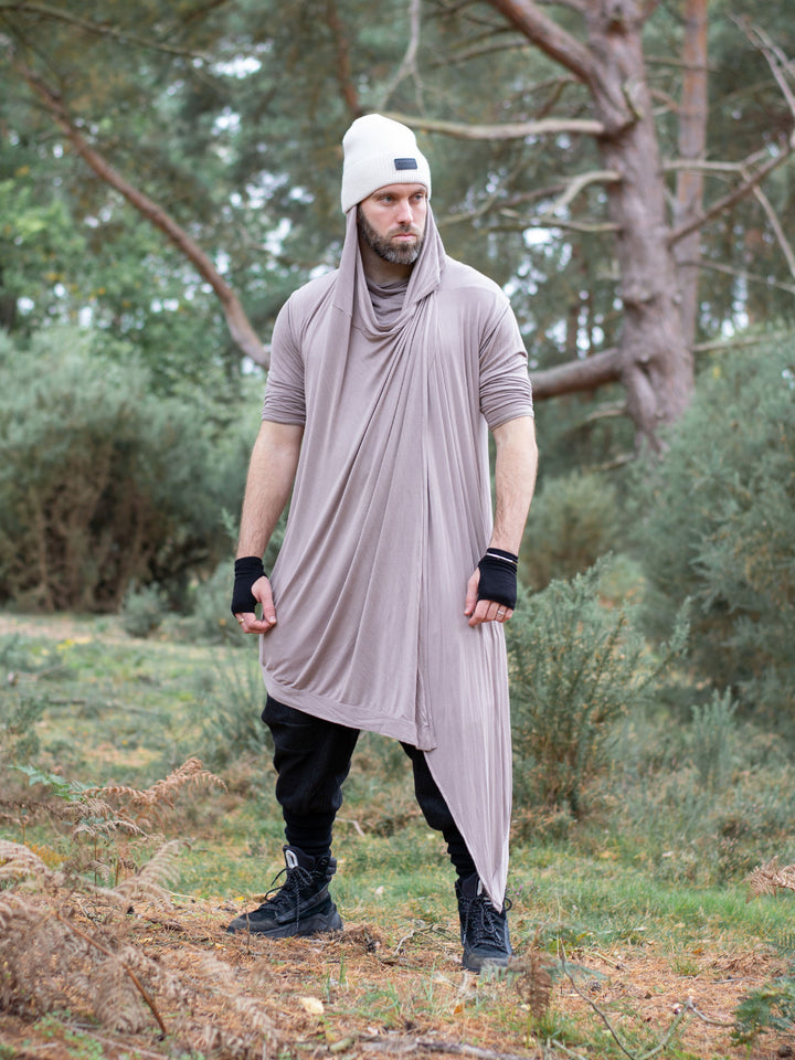 Men’s avant garde hooded cloak in sand with asymmetric wrap-over cascade design. Features bamboo jersey fabric, oversized hood, and wooden shoulder button for layered post-apocalyptic styling.